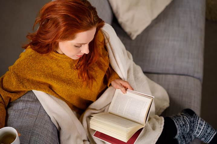 Red haired lady relaxing on a grey couch with a book and a cup of coffee